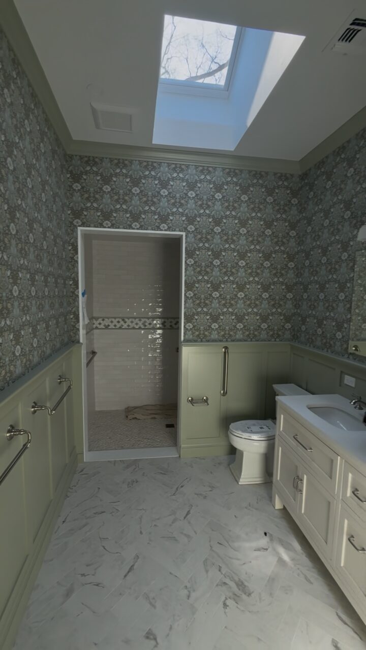 Bathroom with patterned wallpaper, sage green wainscot panelling and a tiled walk-in shower under a skylight.